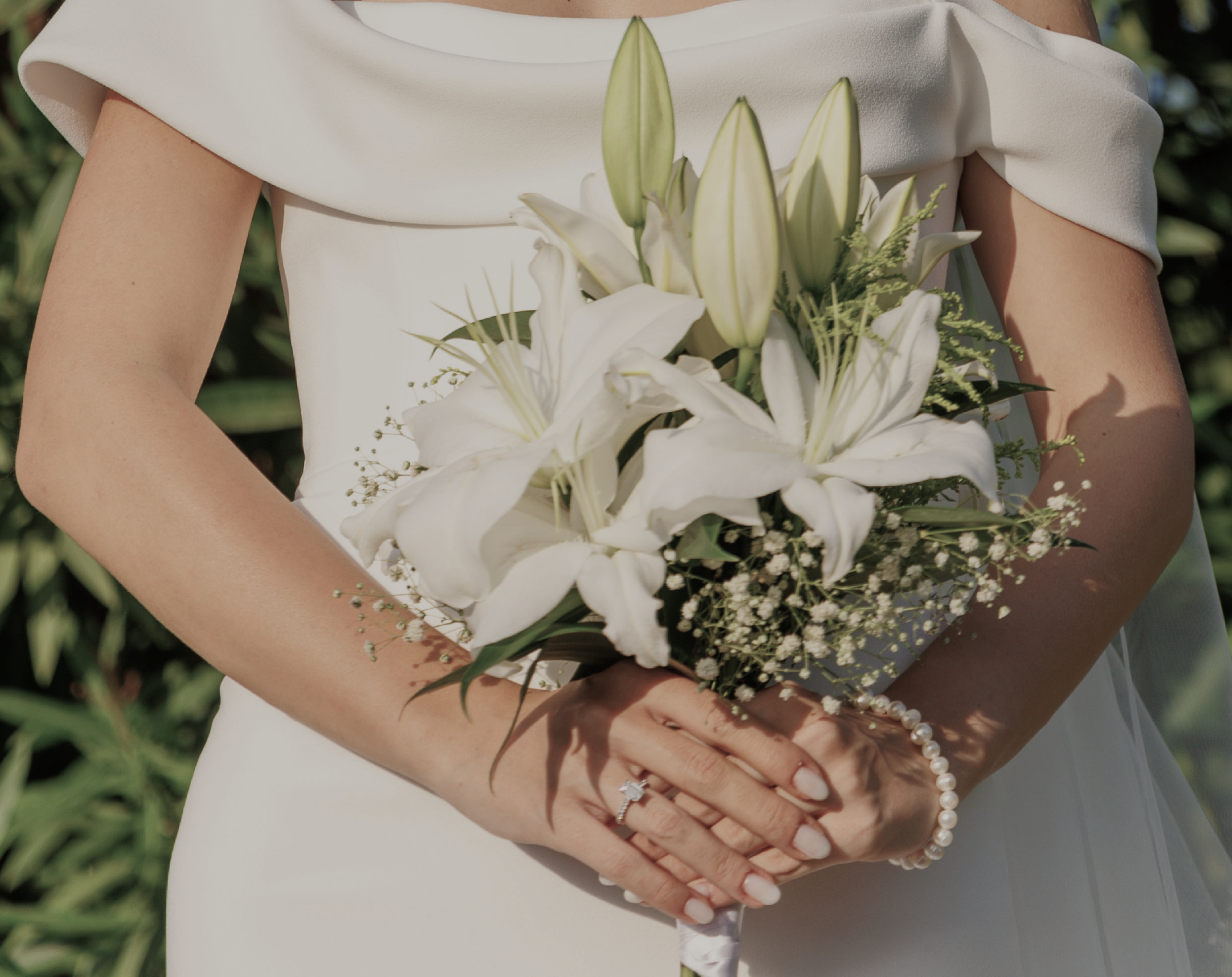 A bride in a white off-the-shoulder wedding dress holding a bouquet of white lilies. Her hands, adorned with a pearl bracelet, delicately clasp the bouquet, drawing attention to her sparkling emerald cut lab-grown diamond engagement ring.