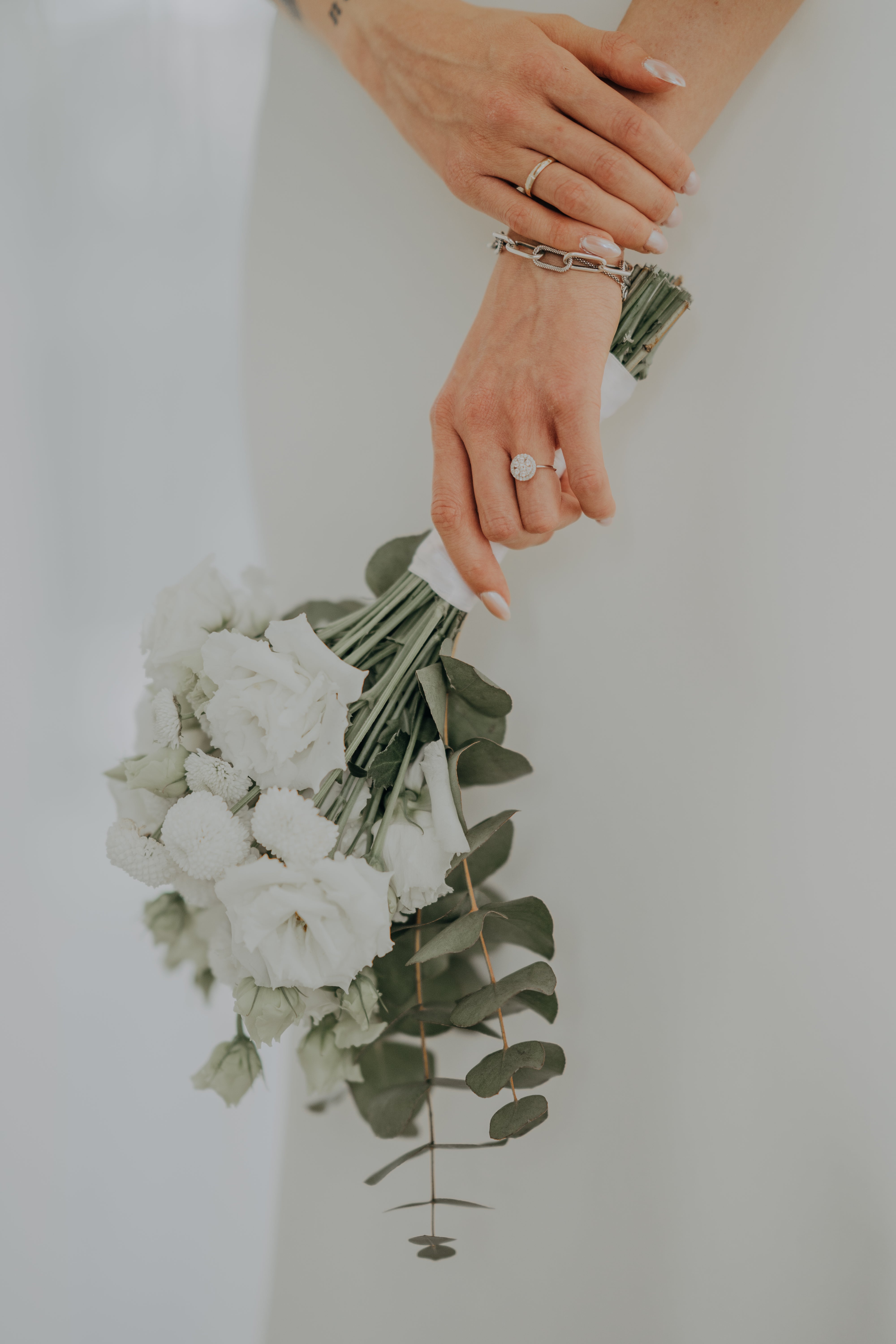 A close-up of a bride’s hands gently holding a bouquet of white flowers and eucalyptus leaves. Her left hand prominently displays a custom-made, round-cut engagement ring with intricate detailing. A gold wedding band and a stylish silver bracelet complement the elegant and personalized bridal look.