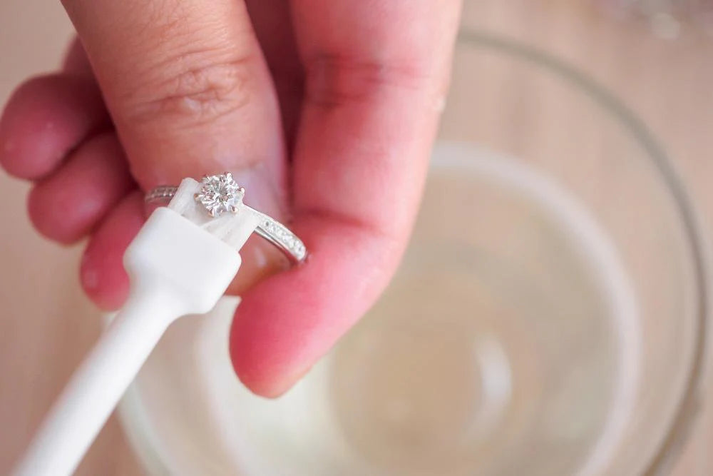Close-up of a hand cleaning a solitaire diamond wedding ring using a small soft brush with jewelry cleaning liquid