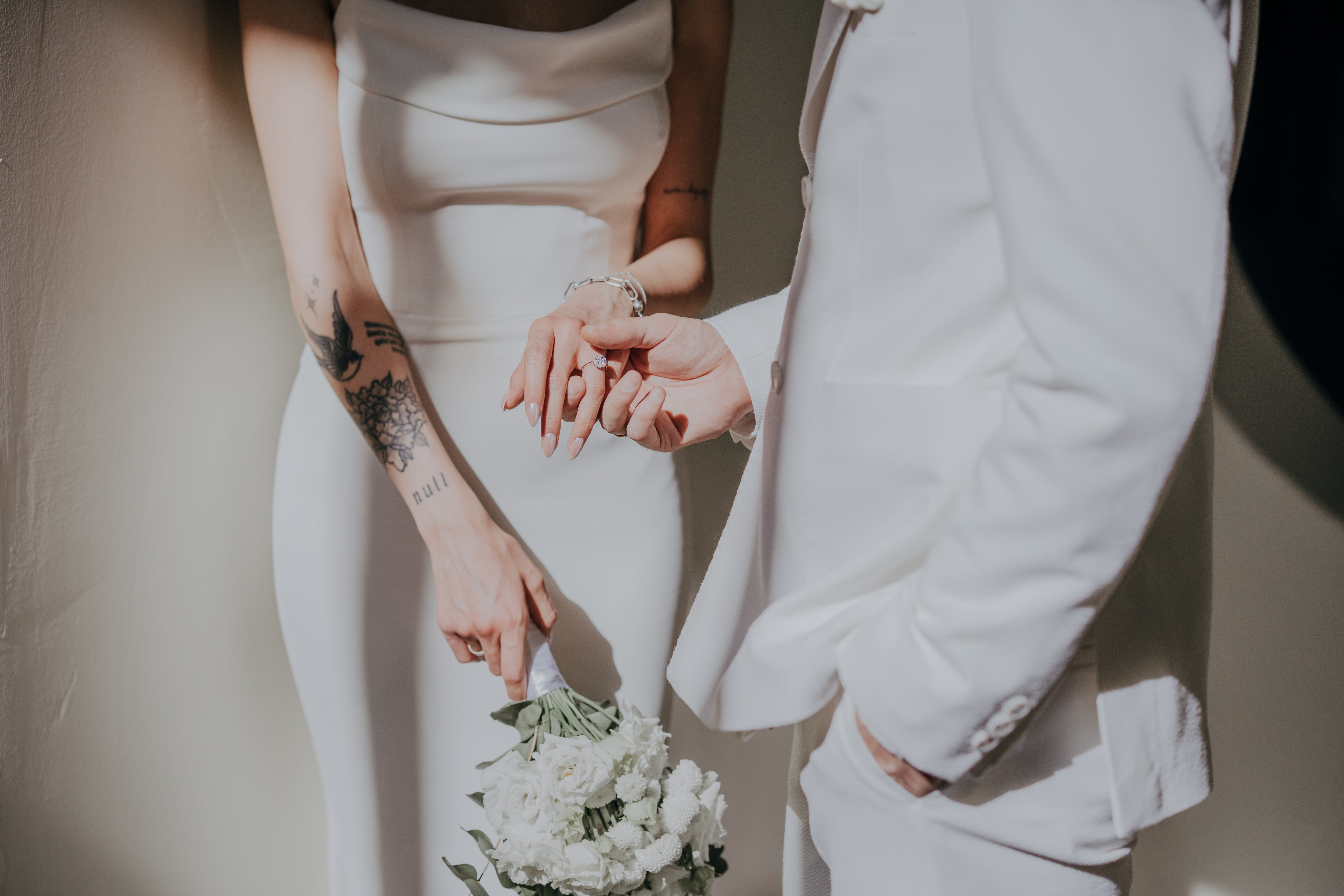 A groom in a white suit gently holds the bride’s hand, admiring her custom-made engagement ring. The bride, dressed in a sleek white gown, holds a bouquet of white flowers in her other hand. Her tattoos add a unique personal touch to the elegant moment, as soft lighting casts a romantic glow.