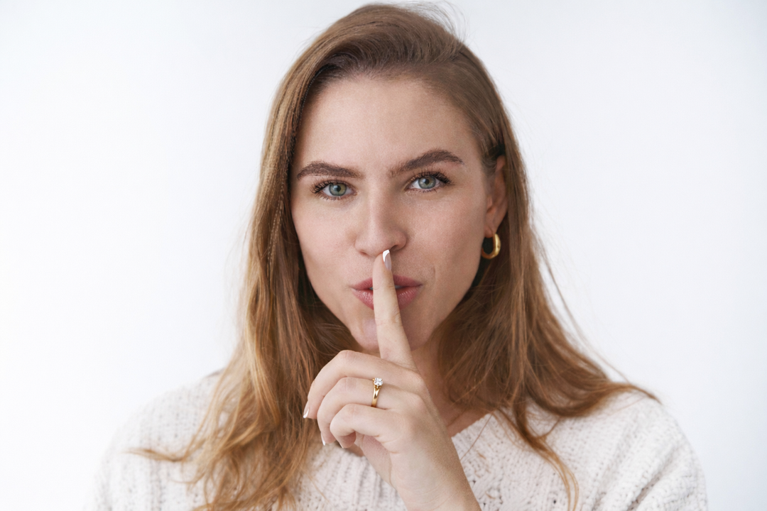A woman with reddish-blonde hair and green eyes looks directly at the camera, making a "shush" gesture with her index finger to her lips. She wears a white knit sweater and a lab-grown diamond ring against a white background.