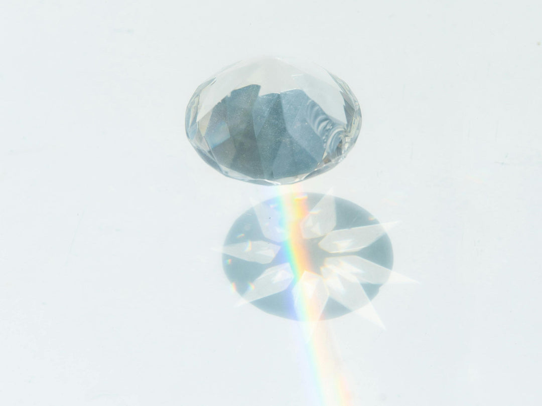 A round-cut diamond on a white surface, creating a subtle rainbow light refraction beneath it that highlights the stone’s brilliance and clarity.